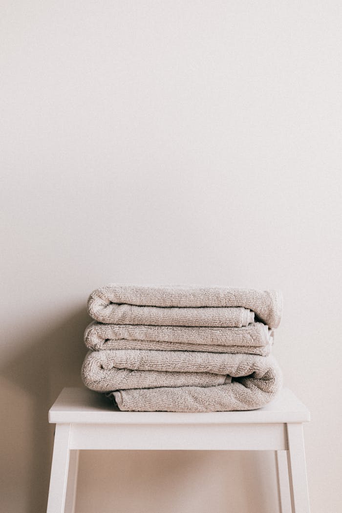 about-us-01 Minimalist composition of white simple stool with clean fresh folded gray towels against beige wall in bathroom